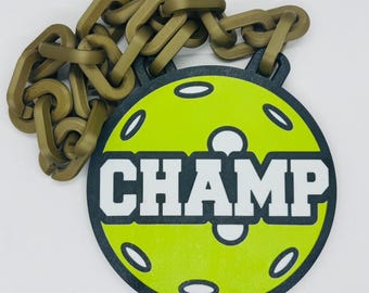 May include: A pickleball-themed medal with a lime green center and the word "CHAMP" in white. The medal is surrounded by a black border and has a series of oval cutouts. A bronze-colored chain is attached to the medal.