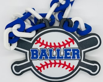 May include: A baseball-themed charm with a white baseball design, red stitching, and the word "BALLER" in blue. The charm is attached to a chain of white and blue links. The charm is a sports-themed accessory.