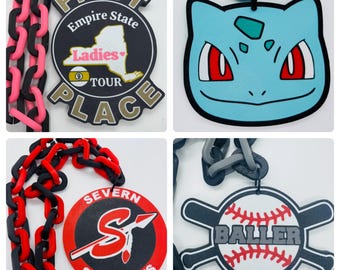 May include: Four different lanyard keychains with various designs. The first keychain features a black and pink chain with a gold and black circular tag that says "First Place" with the words "Empire State Ladies Tour" inside. The second keychain features a blue and gray chain with a blue cartoon character with red eyes. The third keychain features a red and black chain with a black and red circular tag that says "Severn Seminoles" with a red "S" and spear inside. The fourth keychain features a gray chain with a black and white baseball with the word "Baller" on it and two crossed baseball bats.