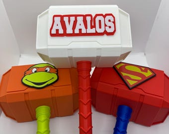 May include: Three toy hammers with different colored handles and tops. The top of the white hammer has the name "AVALOS" in red letters. The orange hammer has a Teenage Mutant Ninja Turtles character on the top. The red hammer has a Superman logo on the top.