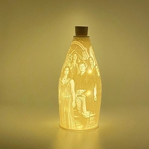 May include: A ceramic bottle lamp with a cork top and a photo of a group of people etched into the side. The lamp is lit and glowing.