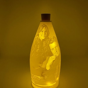 May include: A white ceramic bottle lamp with a cork top. The bottle features a 3D image of a couple. The lamp is lit and glowing.