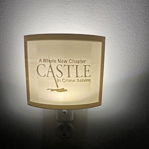 May include: A wooden night light with a curved shape and a white electrical plug. The light features an engraved design that says "A Whole New Chapter CASTLE In Crime Solving" with a drawing of a pen.