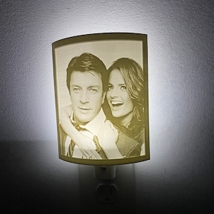May include: A white night light with a curved, translucent plastic shade featuring a black and white photo of a couple. The night light is plugged into a standard electrical outlet.