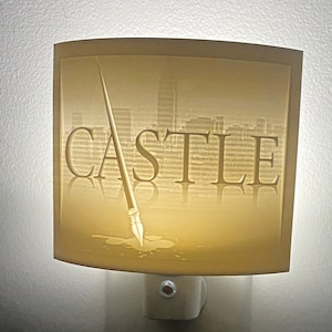 May include: A white night light with a curved, translucent shade. The shade features a cityscape silhouette and the word "CASTLE" in a stylised font. A pen is depicted in the centre of the shade, with ink dripping from the tip.