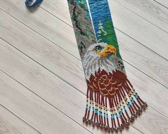May include: A beaded necklace featuring a colourful image of an eagle and a landscape. The necklace has a blue strap and a fringe of beads at the bottom.