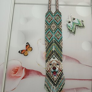 May include: A long beaded necklace with a dog's face in the center. The necklace is made of white, green, blue, and brown beads. The dog's face is white with brown spots. The necklace has a fringe at the bottom. The necklace is paired with matching beaded earrings.