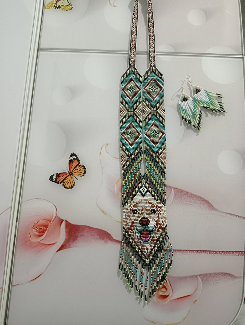 May include: A long beaded necklace with a dog's face in the center. The necklace is made of white, green, blue, and brown beads. The dog's face is white with brown spots. The necklace has a fringe at the bottom. The necklace is paired with matching beaded earrings.