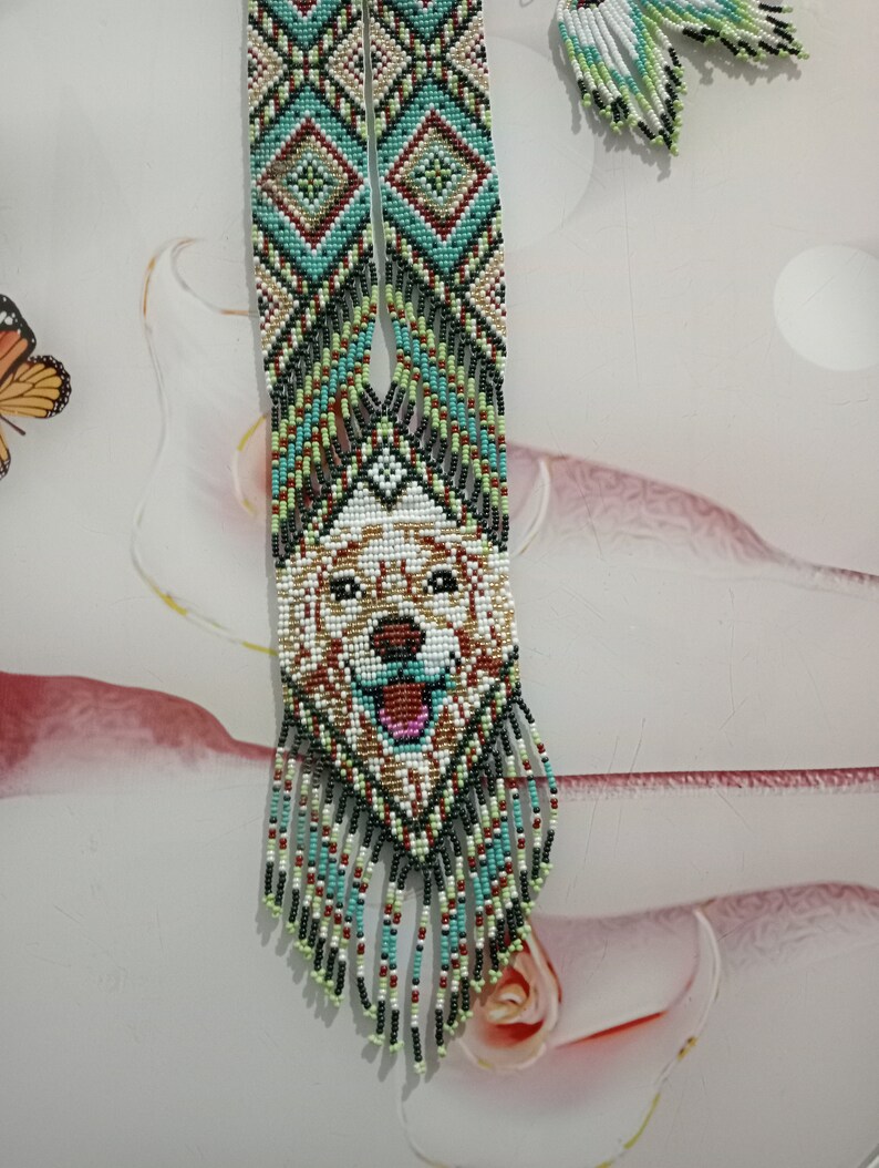 May include: A beaded necklace with a dog face design in the center. The necklace is made with green, blue, white, red, and black beads. The dog face is white with brown and black details. The necklace has a fringe of beads at the bottom.