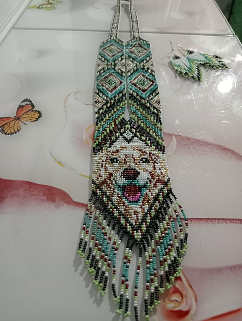 May include: A beaded necklace with a dog's face in the center, surrounded by geometric patterns in shades of green, blue, white, and black. The necklace has long fringe tassels in the same colors. Matching earrings are also included.