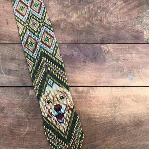 May include: A beaded necklace with a green, yellow, and white geometric pattern. The bottom of the necklace features a beaded image of a golden retriever dog.