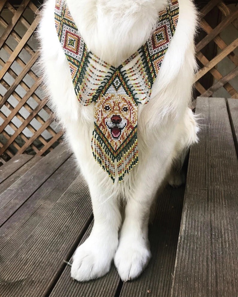 May include: A white dog wearing a beaded necklace with a dog face design. The necklace is green, brown, and gold with a fringe.