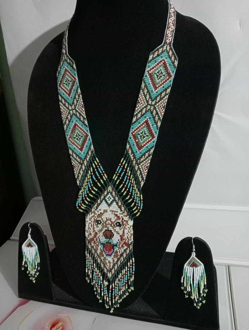 May include: A beaded necklace and earrings set featuring a dog's face in the center of the necklace. The necklace is made of black, white, green, red, and brown beads. The earrings are made of black, white, green, and light blue beads.