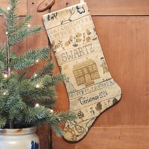 May include: A beige Christmas stocking with embroidered details, including the name "SWARTZ" and "Christmas 2024". The stocking features a house and other decorative elements. A small Christmas tree in a ceramic pot is to the left.