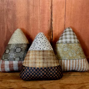 May include: Three decorative fabric cone-shaped objects with a patchwork design. Each cone features different patterns and colors, including polka dots, plaid, and floral prints. The cones are arranged on a wooden surface against a brown wooden background.
