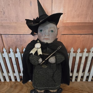 May include: A handmade doll dressed as a witch, wearing a black hat and cape. The doll has a gray knitted dress and holds a needle. A ribbon on the doll says "The Witch". The doll is standing in front of a white picket fence.