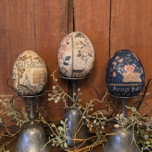 May include: Three embroidered eggs on silver stands, each with a different design. The first egg has the text "Mary Brawn Born Februry" and a small house. The second egg has a floral design. The third egg has the text "I drop her" and a floral design.