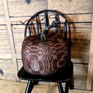 May include: A decorative, burgundy-colored pumpkin with a lace-like pattern and pumpkin illustrations, sits on a black chair. The pumpkin has a dark stem and is a fall-themed decorative item.