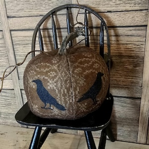 May include: A brown fabric pumpkin with a textured pattern and two dark blue crow silhouettes. The pumpkin sits on a black wooden chair, with a dried vine extending from the top.