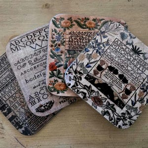 May include: Four square coasters with a vintage alphabet design. The coasters feature a floral pattern with a black and white alphabet design. The coasters are made of a fabric material and have a rustic look.