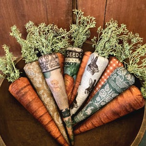 May include: A bowl of eight handmade fabric carrots with different patterns, including a bunny, a seed packet, and a floral design. The carrots are in a wooden bowl with green sprigs on top.