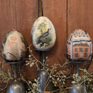 May include: Three decorative eggs on silver stands. The eggs are covered in fabric and feature different designs. One egg has a bird and tree design, another has a house design, and the third has a floral design.