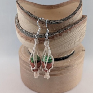 May include: A pair of dangle earrings featuring miniature potted plants. Each earring has a small, terracotta-colored pot holding a green plant, suspended within a macrame-style woven holder. The earrings hang from silver-colored hooks, showcasing a bohemian aesthetic.