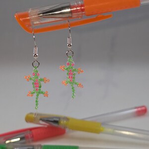 May include: A pair of dangle earrings featuring lizard-shaped charms crafted from small green, pink, and orange beads. The lizard charms are suspended from silver-toned metal earring hooks. The earrings are displayed hanging from an orange pen.