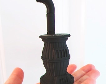 Könnte beinhalten: Ein Miniaturmodell eines schwarzen Ofens mit einem gebogenen Schornstein. Der Ofen hat einen abgerundeten Sockel und einen zylindrischen Körper mit vertikalen Rillen und rechteckigen Lüftungsschlitzen. Der Schornstein erstreckt sich nach oben und biegt sich nach links.