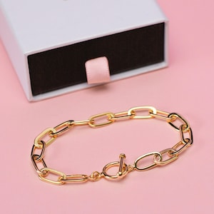 Op de afbeelding: Een gouden kettingarmband met een toggle-sluiting. De armband ligt op een roze achtergrond.