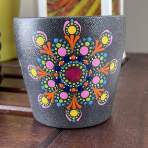 May include: A small, dark gray ceramic pot with a textured surface. The pot is decorated with a colorful mandala design featuring pink, yellow, blue, and orange dots. The mandala is centered on the pot and radiates outwards.