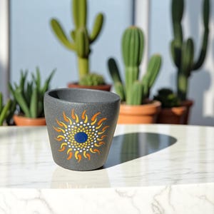 May include: A small gray ceramic planter with a painted design of a sun with orange and yellow rays and a blue center.