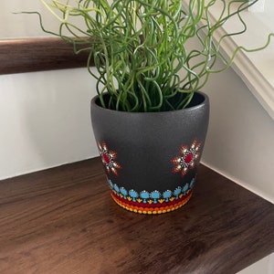 May include: Black ceramic planter with a hand-painted design featuring red, white, and yellow dots forming a flower pattern. The planter is filled with a green plant with long, thin stems.