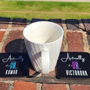 Peut inclure: Deux sous-verres carrés noirs avec du texte blanc qui dit "Actually it's Dr. Kumar" et "Actually it's Dr. Victorova". Les sous-verres sont sur une surface en brique avec une tasse à café blanche.