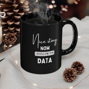 May include: A black ceramic mug with the text "Nice story, NOW SHOW ME THE DATA" in white. The mug is filled with a dark liquid, possibly coffee or tea, and steam rises from the top. The mug has a curved handle.