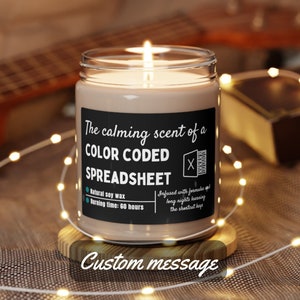 Peut inclure: Une bougie en cire de soja brune avec une étiquette noire qui indique "The calming scent of a COLOR CODED SPREADSHEET". L'étiquette comprend également une petite icône de feuille de calcul avec un X dessus. La bougie a une durée de combustion de 60 heures. La bougie est posée sur une base en bois avec le texte "Custom message" imprimé en dessous.