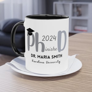 Könnte beinhalten: Weiße Keramik-Kaffeetasse mit schwarzem Rand und Henkel. Die Tasse hat ein graues Design mit der Aufschrift "PhD 2024 inishe" und einem Graduation-Cap-Symbol. Unter dem Design steht "DR. MARIA SMITH Karolina University".