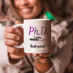 Puede incluir: Taza de cerámica blanca con el texto "Ph.D. loading" en morado y gris, un gráfico de barra de carga y las palabras "keep going!" en negro. La taza tiene un asa blanca.