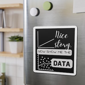 May include: A black and white rectangular magnet with the text "Nice story, NOW SHOW ME THE DATA." The magnet features a graph and a data chart design. The magnet is attached to a silver fridge door with two round magnets.