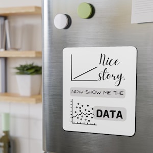 Puede incluir: Un imán rectangular blanco con el texto "Nice story. Now show me the DATA" en negro. El imán presenta dos gráficos de líneas negras. El imán está adherido a la puerta plateada de un frigorífico con dos imanes redondos.