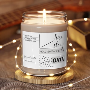 以下が含まれることがあります： 白いラベルに「Nice story. Now show me the data.」と書かれた茶色のキャンドル。ラベルには折れ線グラフと小さな円グラフも含まれています。キャンドルは木製のコースターの上に置かれています。