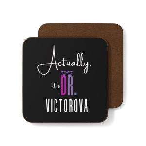Peut inclure: Sous-verre carré noir avec le texte "Actually, it's DR. VICTOROVA" en blanc et violet. Le dessous de verre a une bordure marron et est posé sur un autre dessous de verre avec une surface marron.