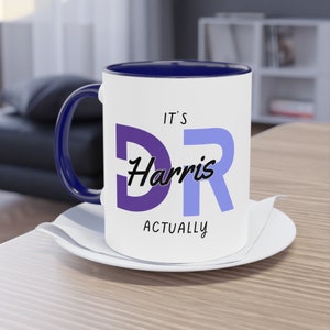 Pode incluir: Caneca de café branca com borda e alça azuis. Tem o texto "It's Dr. Harris Actually" impresso em preto com um "D" roxo e um "R" azul.