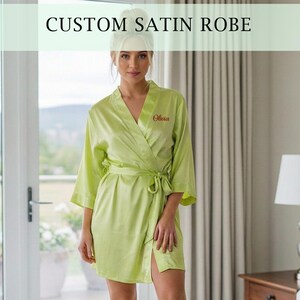 May include: A light green satin robe with the text "CUSTOM SATIN ROBE" at the top. The robe has a wrap-around design with a matching belt. The name "Olivia" is embroidered on the front. The robe is knee-length.