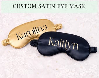 Pode incluir: Duas máscaras de olhos de cetim, uma dourada e outra azul marinho, são exibidas. A máscara dourada tem o nome "Karolina" impresso, enquanto a máscara azul marinho tem "Kaitlyn" impresso. O texto "CUSTOM SATIN EYE MASK" está no topo.