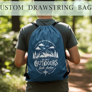 Può includere: Una borsa blu con coulisse e cordino nero è indossata sulla schiena di una persona. La borsa presenta una grafica bianca di montagne, alberi, nuvole e le parole "The Great Outdoors". Il testo "Custom Drawstring Bags" è in alto.