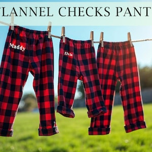 May include: Two pairs of red and black plaid flannel pajama pants hanging on a clothesline. One pair has the name "Maddy" embroidered on the front and the other pair has the name "Elvis" embroidered on the front.