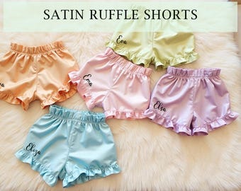 May include: Five pairs of satin ruffle shorts in different colors, each with a name embroidered on the front. The colors are peach, pink, lavender, light blue, and light green.