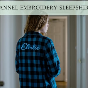 Könnte beinhalten: Eine Person trägt ein blau-schwarz kariertes Flanell-Nachthemd mit dem Namen "Elodie" in hellblauer Schrift auf dem Rücken gestickt. Das Bild enthält auch den Text "FLANNEL EMBROIDERY SLEEPSHIRTS".