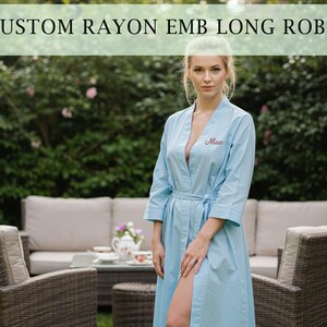 Könnte beinhalten: Hellblaue, bestickte, lange Robe aus Rayon mit dem Namen "Mae" auf der Brust. Die Robe hat einen Bindegürtel und Dreiviertelärmel. Das Bild zeigt die Robe im Freien mit Garten und Terrassenmöbeln im Hintergrund.